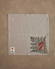 Coral & Tusk - Mushrooms and Ferns Bouquet Dinner Napkin with embroidered botanical motif, featuring mushrooms and ferns, on a brown surface.