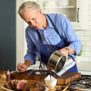Middle-aged man pours gravy from Hestan | Cookware, 1.5qt Saucepan + Lid over roast beef, showcasing its precision pouring feature in a home kitchen setting.