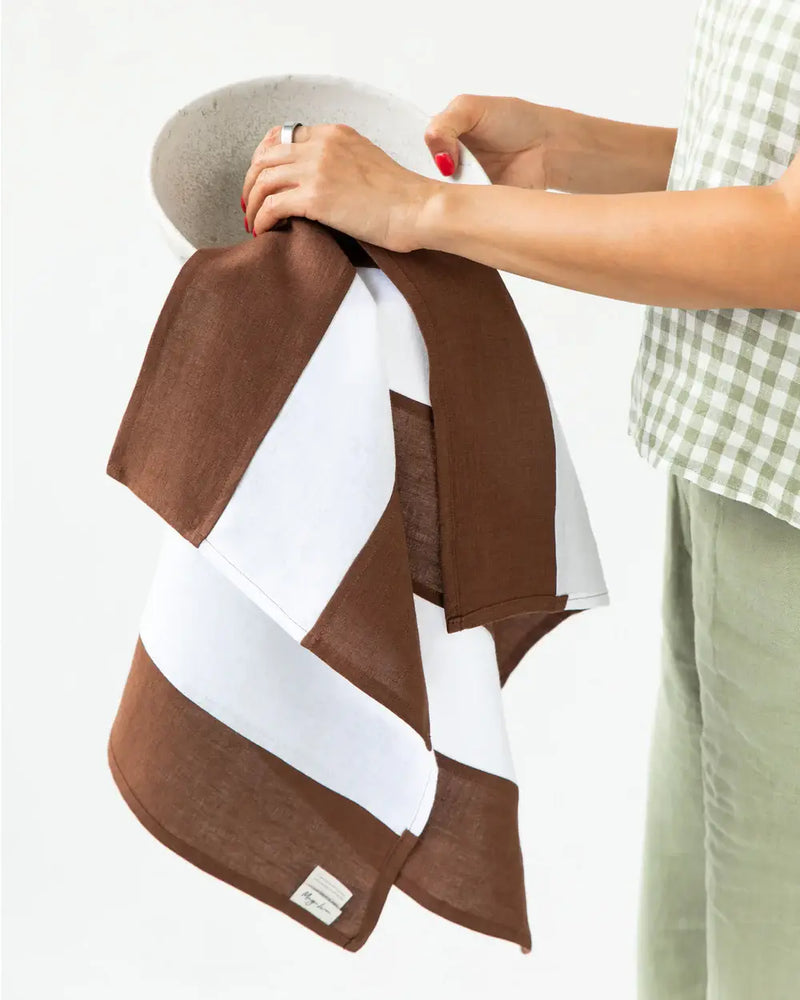 Person holding MagicLinen's striped linen tea towel, chocolate brown, part of zero-waste initiative, featuring a hanging loop, ideal for stylish and eco-friendly kitchen use.