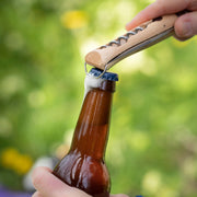 A person uses the Opinel | Nomad Cooking Kit's corkscrew knife to open a beer bottle outdoors, showcasing its functionality for outdoor culinary adventures.