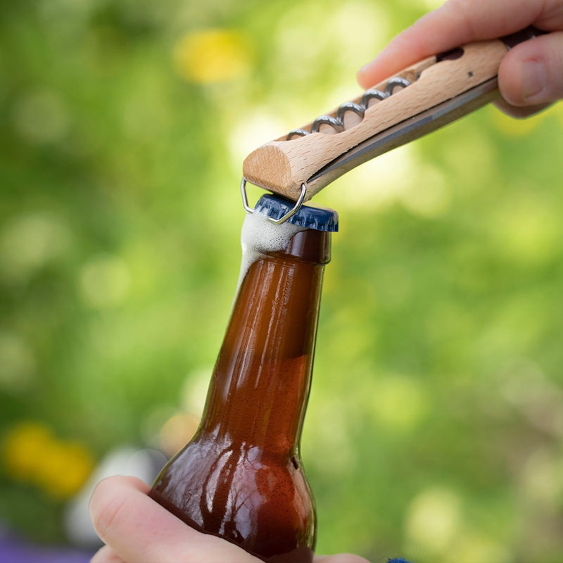A person uses the Opinel | Nomad Cooking Kit's corkscrew knife to open a beer bottle outdoors, showcasing its functionality for outdoor culinary adventures.