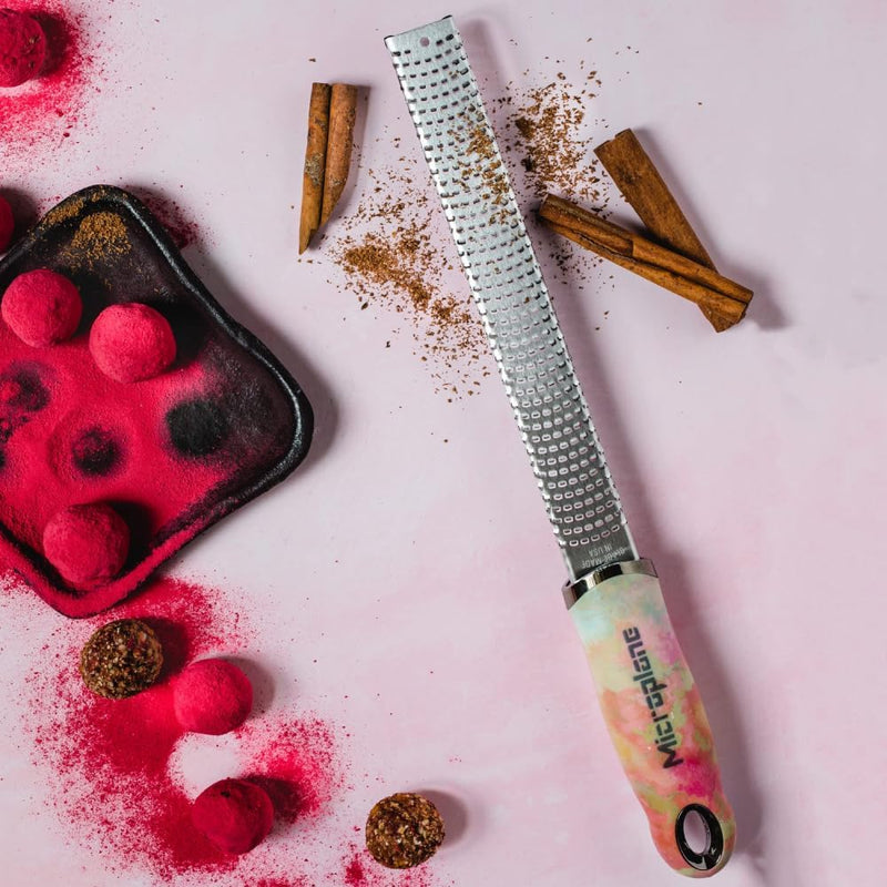 Microplane | Premium Zester, Grater - Ombre on a pink surface, surrounded by cinnamon sticks, pink-coated truffles, and a small black tray with more truffles.