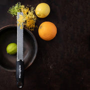 Microplane | Premium Zester, Grater - Black shown over a wooden bowl with a lime, surrounded by a lemon, orange, and piles of citrus zest.