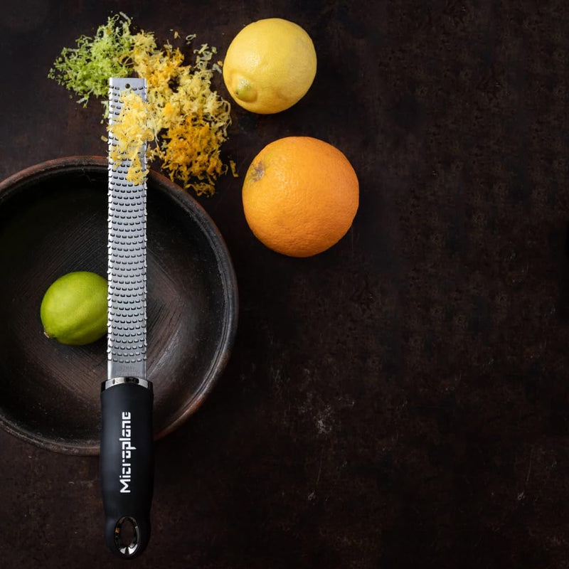 Microplane | Premium Zester, Grater - Black shown over a wooden bowl with a lime, surrounded by a lemon, orange, and piles of citrus zest.
