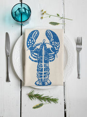 Hearth and Harrow Lobster Tea Towel, organic cotton with blue-violet lobster design, displayed on a white table setting with silverware and plate.