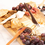 Wooden spreader by Collin Garrity elegantly spreads soft cheese on charcuterie board with grapes, bread, crackers, firmer cheese, cured meat, and fruit jam.
