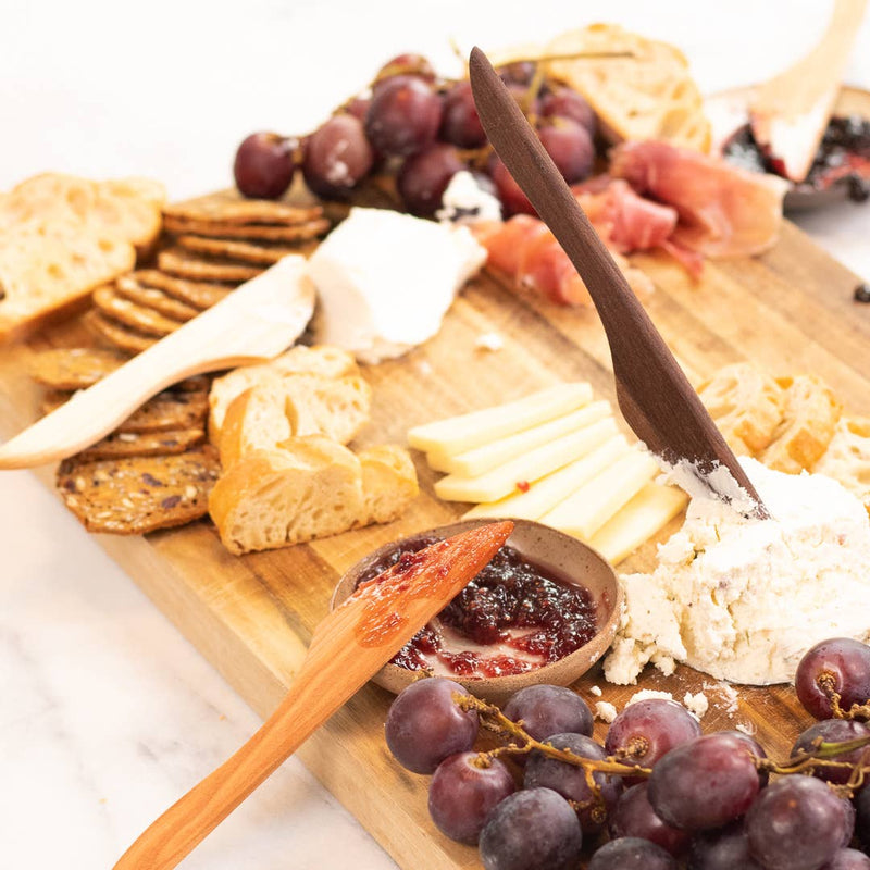 Wooden spreader by Collin Garrity elegantly spreads soft cheese on charcuterie board with grapes, bread, crackers, firmer cheese, cured meat, and fruit jam.