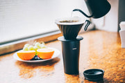 HARIO USA - V60 Metal Dripper: Silver in action, brewing coffee over a travel mug with gooseneck kettle; fresh fruit on wooden table.