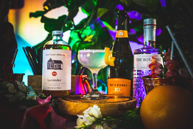 Pink House Alchemy - Lavender Simple Syrup bottle with surrounding bottles and a glass, showcasing our aromatic syrup ideal for cocktails and culinary creations.