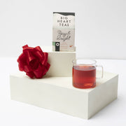 Big Heart Tea Co. Rosy & Bright 10 ct Tea Bags with a glass of brewed magenta tea, accompanied by a festive red bow, on white pedestals.