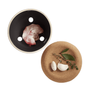 Emile Henry | Garlic Pot, Burgundy: A ceramic dish with ventilation holes showcasing fresh garlic, accompanied by a cork trivet with bay leaves and thyme.