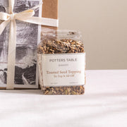 Potters Table - Top it All Off: Seeded Topping in a clear bag, perfect for adding crunch and flavor to meals, sits beside a kraft-paper gift box.