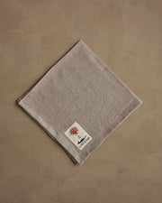 Coral & Tusk - Pressed Leaves Dinner Napkin featuring autumnal leaf designs, acorns, and quail feathers, with a small label, laid on a textured surface.