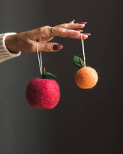 Hand with gold ring holds two Farmhouse Pottery - Felted Fruit Ornaments: Clementine and apple, showcasing handcrafted wool design.