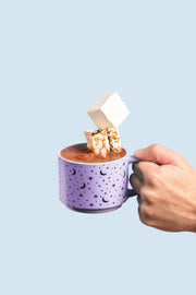 Hand holding a pastel purple mug filled with hot chocolate, topped with two Fluffy Nutter marshmallows—one plain, one with hazelnut crunch.