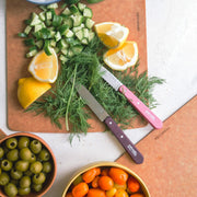 Opinel | Essential Serrated Knife, Pink, displayed on cutting boards with diced cucumber, fresh dill, lemon wedges, surrounded by bowls of olives and grape tomatoes.