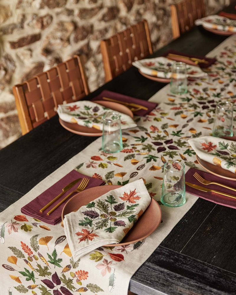 Coral & Tusk - Pressed Leaves Dinner Napkin elegantly folded on a muted pink plate, set on a rustic table with fall-themed decor and gold flatware.