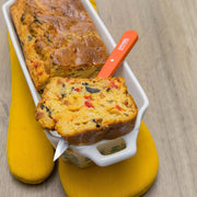 Opinel | Essential Paring Knife, Tangerine, lifting a savory loaf slice with red pepper, black olives, and cheese from a ceramic pan on a wood surface.