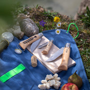 Opinel | Nomad Cooking Kit: Outdoor picnic setup with serrated knife, corkscrew, and peeler on a cutting board, surrounded by cheese, fruits, and flowers.