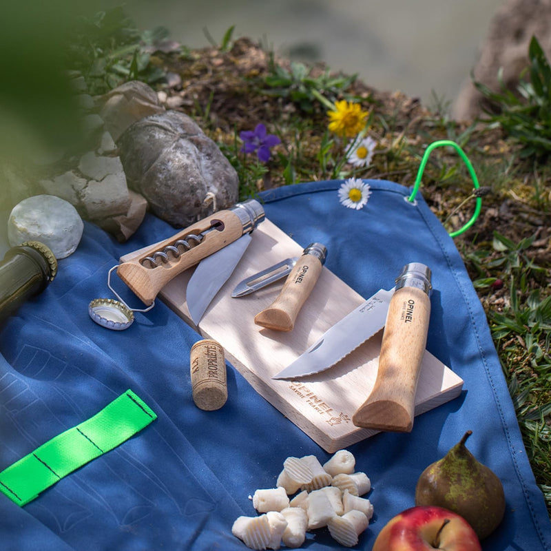 Opinel | Nomad Cooking Kit: Outdoor picnic setup with serrated knife, corkscrew, and peeler on a cutting board, surrounded by cheese, fruits, and flowers.