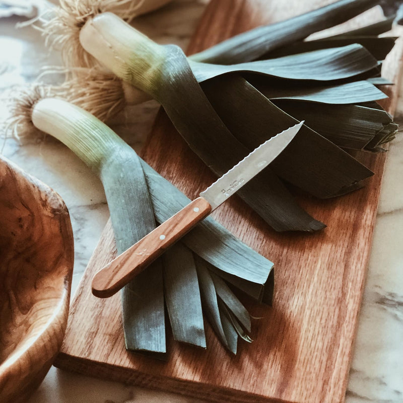 Opinel | Essential Serrated Knife, Pink resting on whole leeks on a wooden board, showcasing its durable beech wood handle and efficient, serrated cutting edge.