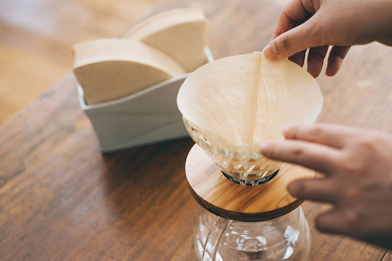 Hands fitting paper filter into glass dripper on wood-topped carafe, with storage box nearby. HARIO USA - V60 Paper Filters: White / 100 Sheets Boxed.