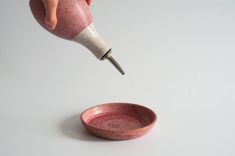 Hand holding RachaelPots - Garlic Scraper: Coral, a two-tone ceramic bottle with pour spout, above a matching dish, ideal for creating garlic puree quickly.