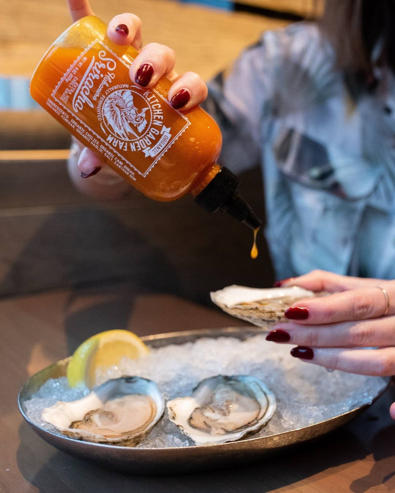 Person drizzles Kitchen Garden Farm - Organic Habanero Sriracha onto a raw oyster on a platter, emphasizing the spicy, fermented sauce's organic ingredients.