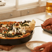 Emile Henry | Chicago Deep Dish Pan, Charcoal: A slice of deep-dish pizza with sausage and greens lifted by a spatula, showcasing the pan's golden crust and cheese stretch.