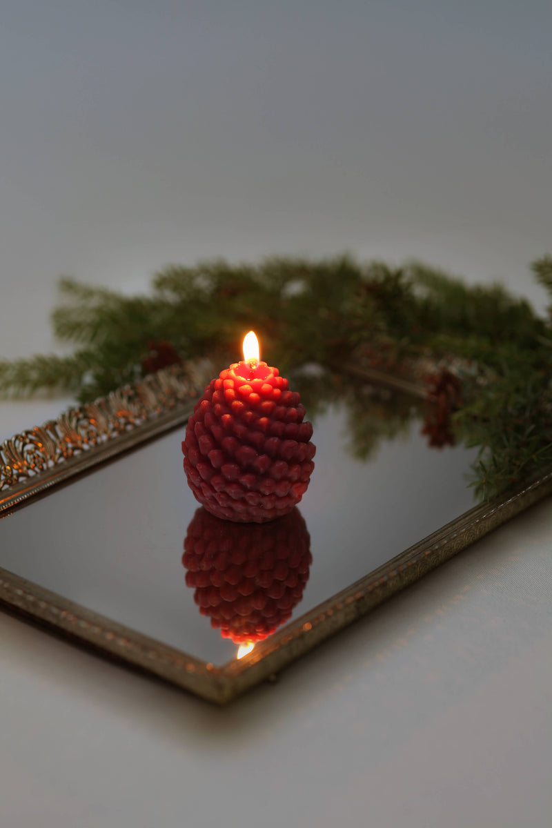 Mo&Co Home Exclusive: Sculpted Beeswax Pine Cone candle on a mirrored tray, surrounded by evergreen sprigs, capturing a holiday ambiance.