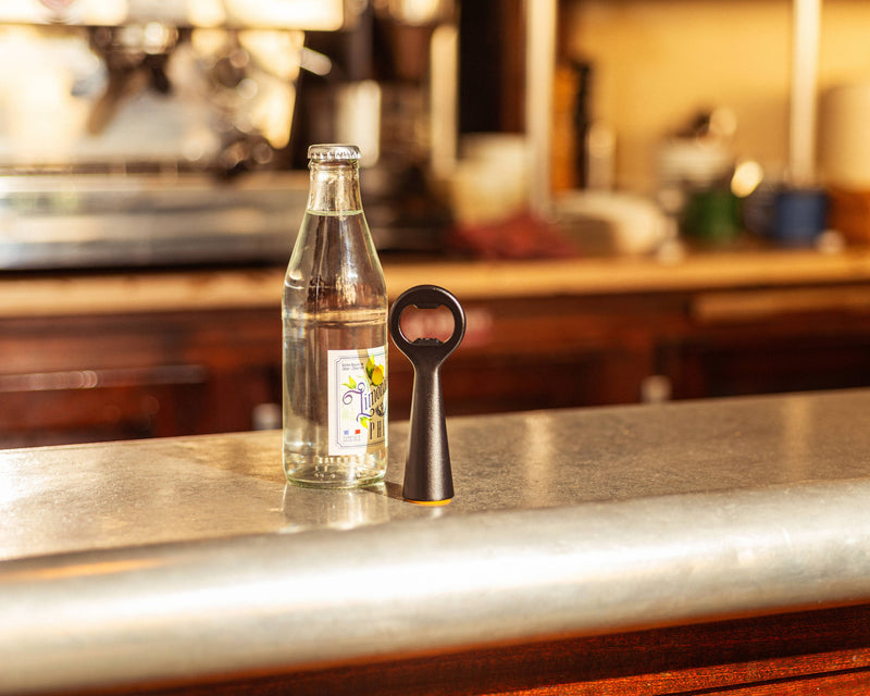 L'Atelier du Vin - USA - Bottle Opener - LE BRASSEUR – BLACK on a metal countertop next to a clear glass bottle of carbonated beverage.
