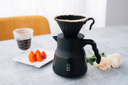 HARIO USA - V60 Metal Dripper: Silver with coffee server and brewed coffee on a tabletop, accompanied by strawberries and roses, highlighting precision brewing in an elegant setup.