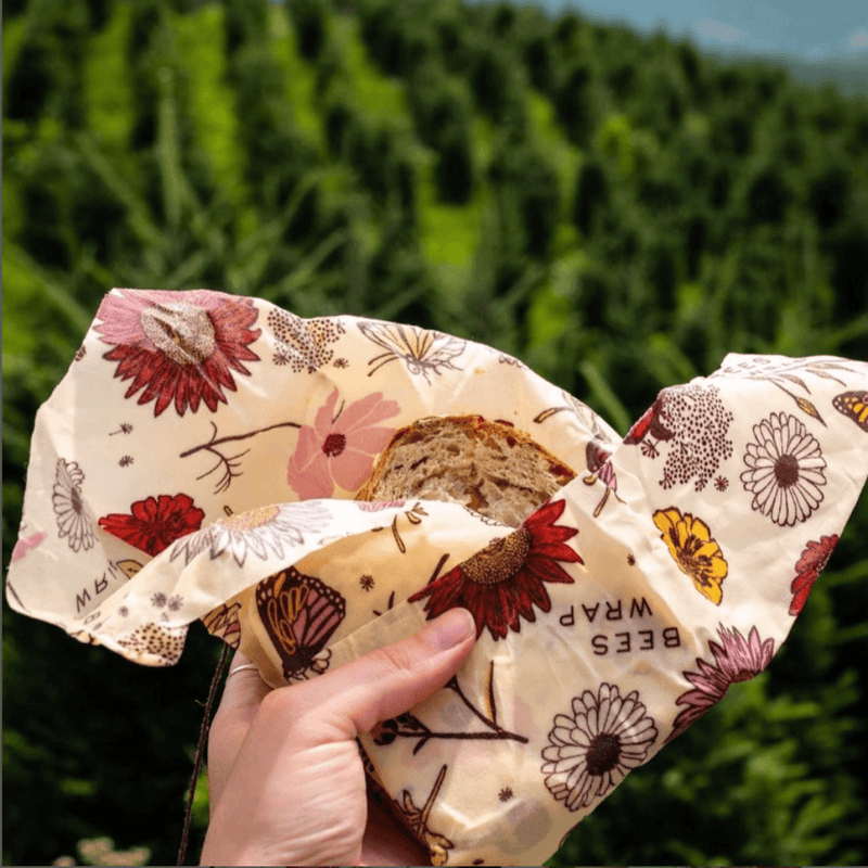 Hand holding a Bee's Wrap - Sandwich Wrap - Meadow Magic, Vegan Wax, covering a sliced loaf outdoors, showcasing its reusable, eco-friendly design with floral and butterfly prints.