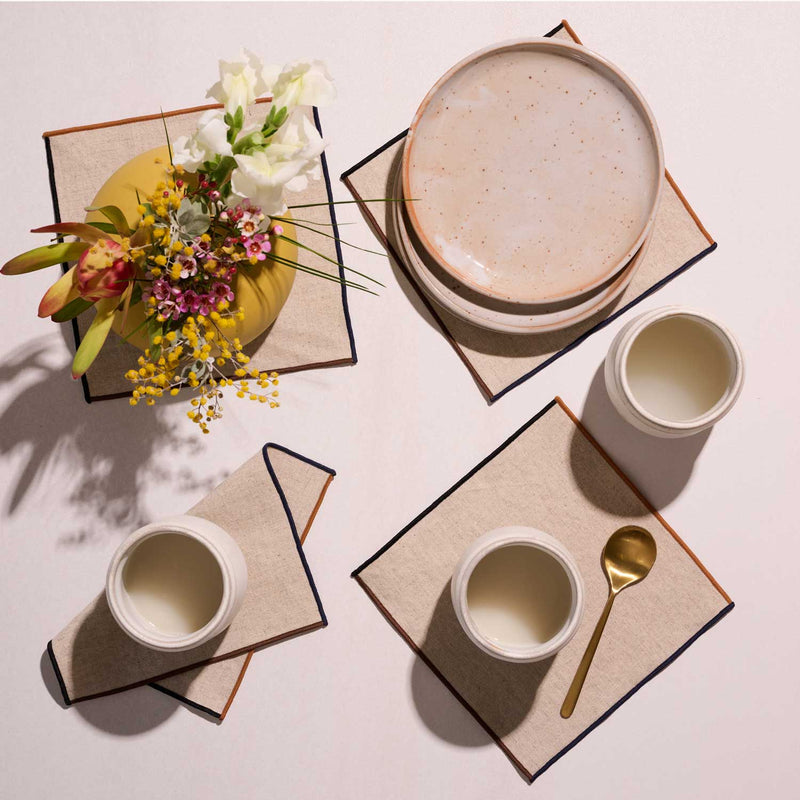 ATELIER SAUCIER - Dune Appetizer Napkins | Set of 4 elegantly complement the tabletop, featuring color-blocked trim, alongside a ceramic plate, vase, cups, and teaspoon.