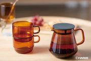 HARIO USA - HARIO Colors Glass Coffee and Tea Server, 600ml: Amber, displayed with stackable mugs, on a wood surface.