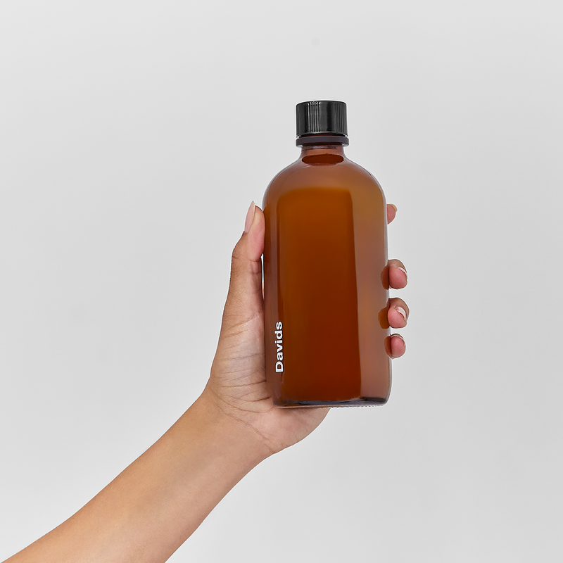 Hand holding Davids Hydroxi™ fresh breath mouthwash refill concentrate in amber-brown glass bottle, highlighting its sustainable, fluoride-free formulation with premium mint oil blend.
