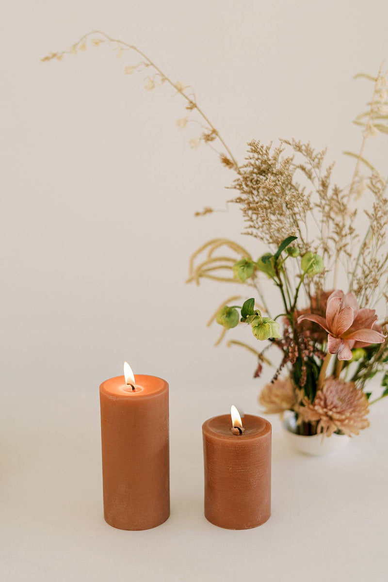 Mo&Co Home - 100% Beeswax Pillar Candles | Terra Cotta: Two candles with soft glow, surrounded by flowers, ideal for home decor and special occasions.