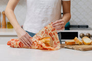 Person wrapping baguette in Bee's Wrap - Bread Wrap - Mushroom Magic, Limited Edition; wooden board with bread slices, kitchen background visible.