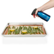 Hand holding Evo Stainless Steel Oil Sprayers - Purple, spraying oil over asparagus on a tray, showcasing precise, non-aerosol distribution for healthier cooking.