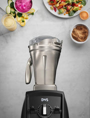 Vitamix | 48 oz Stainless Steel Container shown with a broken lid, surrounded by various food ingredients, highlighting its versatile blending capability.