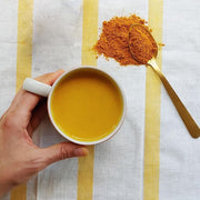 Hand holding a white mug of Spicewalla Golden Milk with turmeric powder and a gold-handled spoon on a striped cloth, emphasizing antioxidant-rich ingredients.