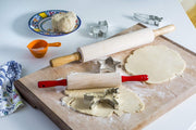 Kid's rolling pin with red handles on a cookie-making setup, featuring rolled dough, metal cookie cutters, and a measuring cup. Perfect for young bakers at Athens Cooks.