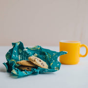 Bee's Wrap - Assorted 3 Pack - Oceans shown with a cookie on a blue cloth beside a yellow mug, highlighting reusable food storage wraps.
