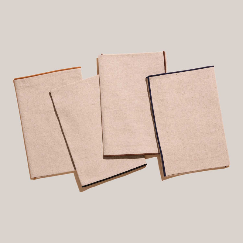 ATELIER SAUCIER - Dune Napkins | Set of 4: Natural linen napkins with bold, color-trimmed edges, neatly stacked, embodying understated elegance for a chic dining experience.