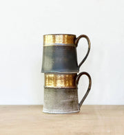 Two stacked Jessica Wertz Ceramics - Black + Gold Mugs with metallic-gold rims and matte grayish bodies on a wooden surface. Each mug holds 12 ounces and is dishwasher safe.