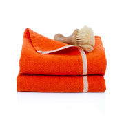 Caravan Home | Chunky Linen Orange Towels feature two folded towels with a white stripe, topped by a small wooden-handled scrub brush, embodying rustic French charm.