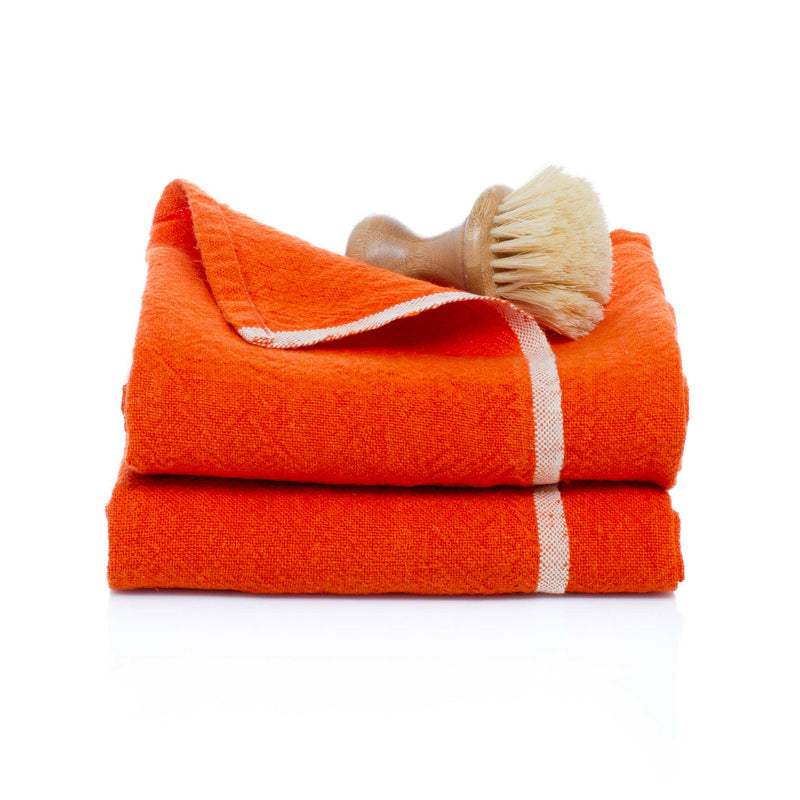 Caravan Home | Chunky Linen Orange Towels feature bright-orange linen with a white border, stacked with a wooden-handled brush, offering a modern rustic aesthetic.