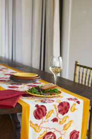 Pomegranate Yellow/Red Table Runner on a rustic dining table, adorned with a salad bowl, plates, and a wine glass, creating a vibrant setting.