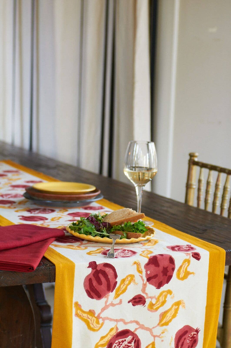 Pomegranate Yellow/Red Table Runner on a rustic dining table, adorned with a salad bowl, plates, and a wine glass, creating a vibrant setting.