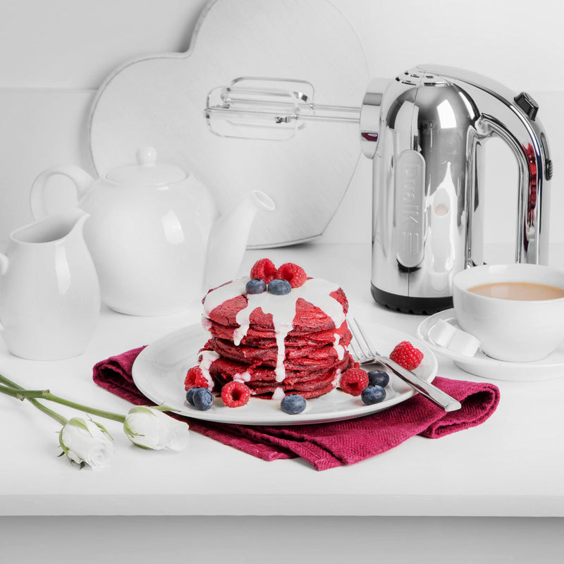 Dualit Hand Mixer in polished chrome stands beside red-velvet pancakes with cream and berries, showcasing its sleek design amidst a vibrant breakfast setting.